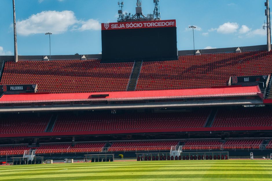 Empty football stadium with red stands, scoreboard, and lush green field under a bright sky.
