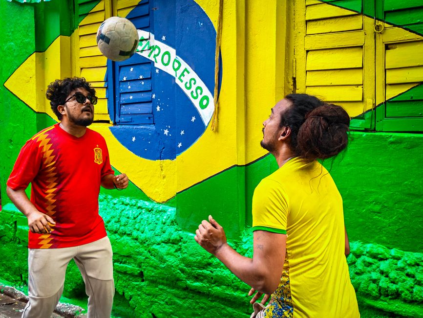 Two men playing soccer in front of a vivid Brazilian flag mural.