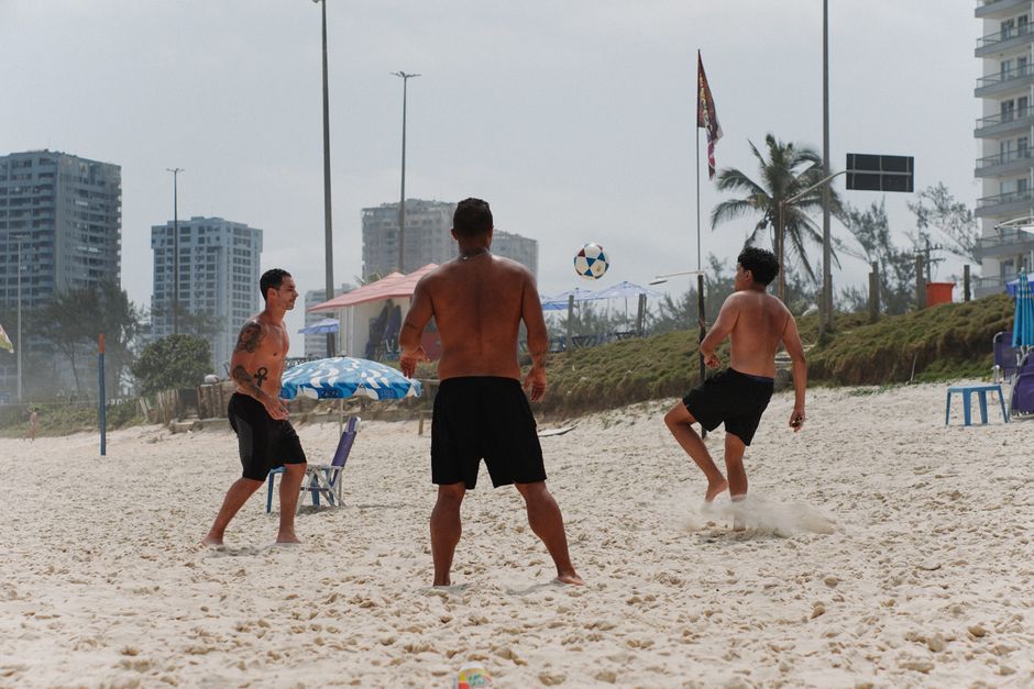 Three men playing soccer on a sandy beach with city skyline and palm trees.