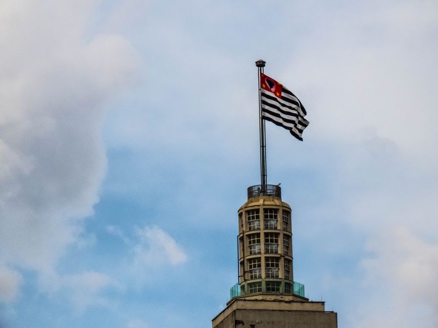 View of Altino Arantes Building in São Paulo with the state flag on top against a blue sky.