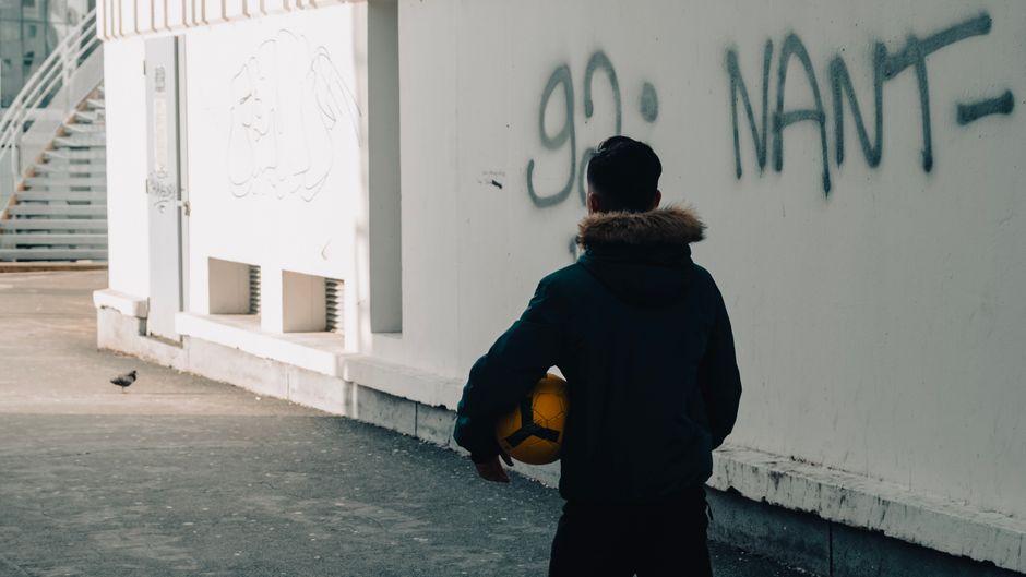 Man stands in urban street holding ball, facing graffiti wall.